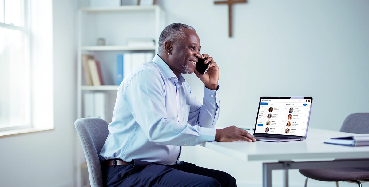 A pastor seated in a bright church office making a follow-up phone call while reviewing member records on a laptop, representing proactive summer re-engagement before families drift away